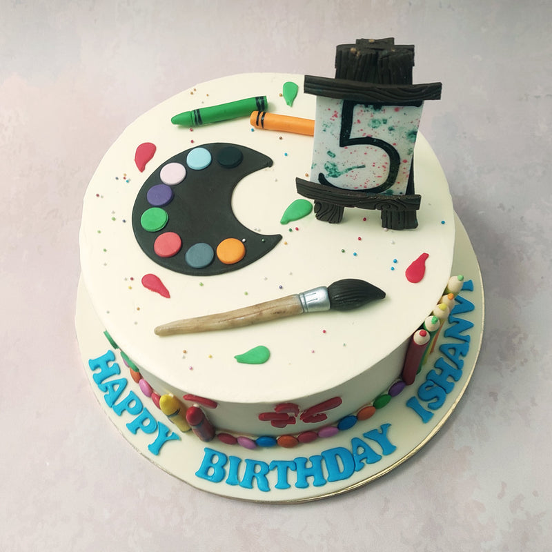 With a white base, like a canvas, this drawing cake showcases a number of artistic icons from realistic crayons engulfing the circumference to an easel and canvas on top.