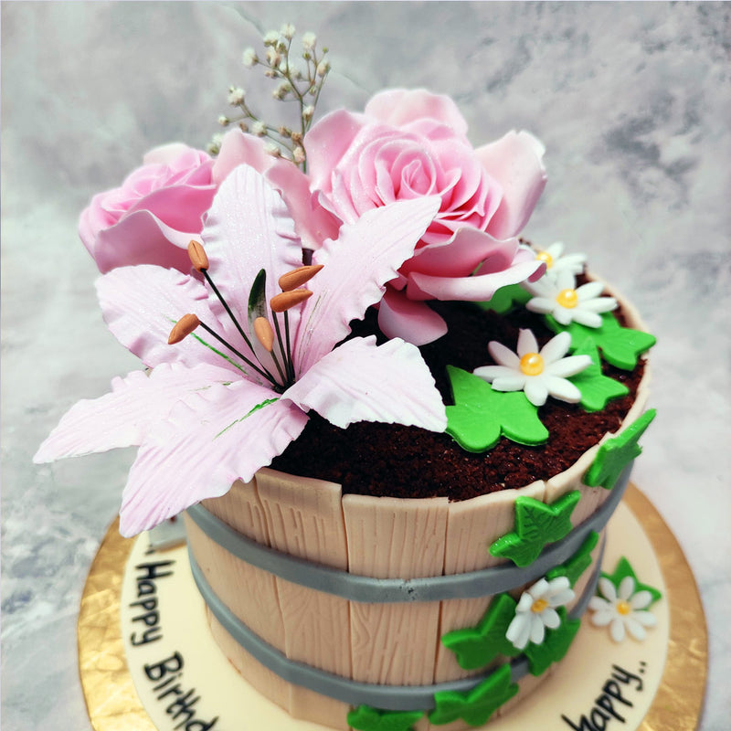 This flower pot cake design features a pot made of textured wooden slabs bound together with two rings of cement. Mud made of cake comprises the inside of the flower cake's pot from which two big, beautiful pink roses bloom alongside a pink lily.