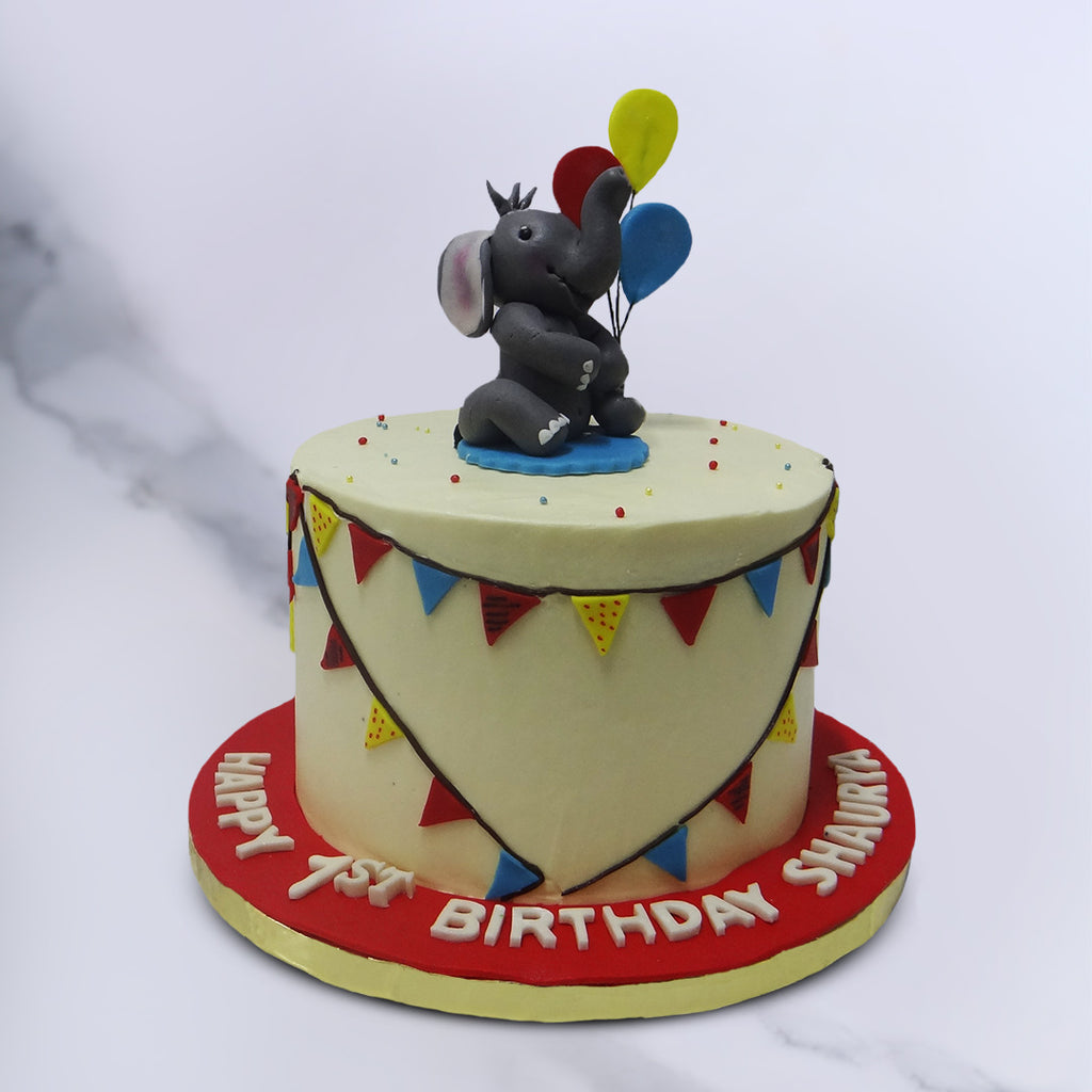 The star edible elephant on this elephant theme cake is sitting on his haunches, holding red ,blue and yellow balloons.