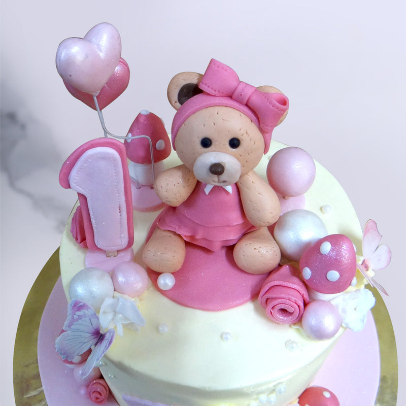 Top view of pink teddy bear cake with balloons and a number 1 figurine on top. This first birthday cake for girls has a cute little teddy bear also