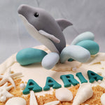 The base of this dolphin cake is a neutral off-white colour with cookie crumbs that represent the sand on a beach and is ornamented with white chocolate seashells and features a dolphin emerging out of it with blue water splashes all around it.