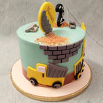 This excavator theme cake has a sky blue base with brick red smears at the bottom. Grey bricks and a yellow excavator decorate the circumference, digging out cookie crumbs that resemble sand.