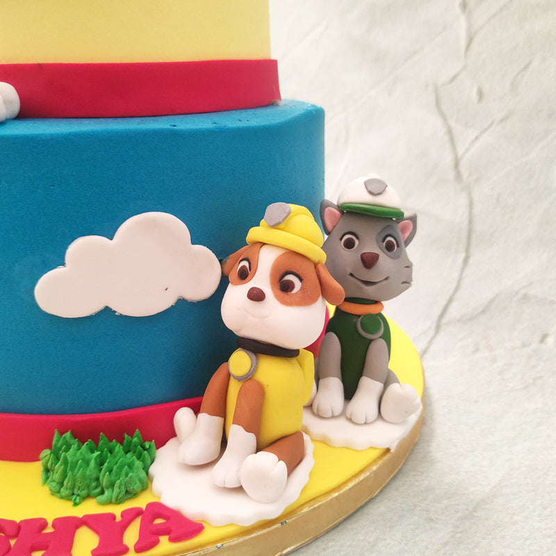 The yellow top tier of this Paw Patrol two tier cake proudly displays the show's logo with detailing done in the form of fluffy white clouds passing by and a collar at the base. Lookout also for the iconic Lookout Tower! 