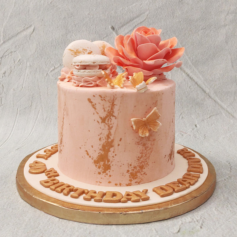 This peach rose cake comes with a tall base, embossed with gold leaf flakes and orange tinted butterflies. On top, in decadent and silky buttercream, spot a giant peach rose highlighting the floral theme of this rose macaron cake. 