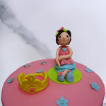 This is a unique take on a kids birthday cake as the star Princess sits on a lilypad beside her crown in a pink dress. Flowers adorn her shoes and leaves adorn her hair and the gold crown that awaits her floats by on another lilipad.