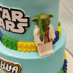 A cartoon, lego-like figurine of Yoda rests on top of the first tier protecting the top tier where similar figurines of Luke Skywalker and Darth Vader are displayed holding blue and white lightsabers like guardians of this two tier Star Wars cake galaxy.