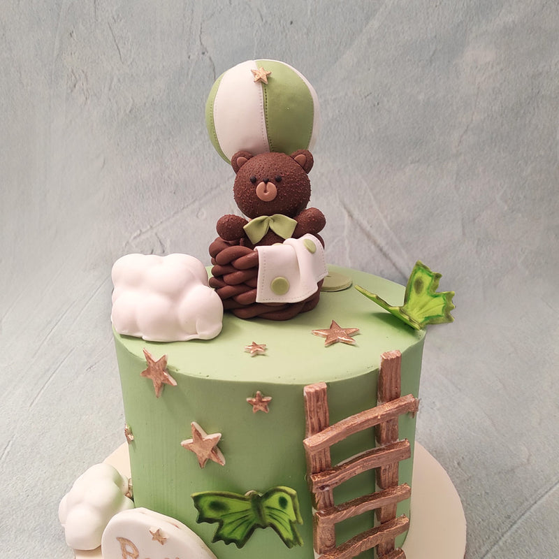 The base of this teddy bear cake comes in a mint green shade with a little model of a  wooden ladder leading up to a small space on top of the teddy bear birthday cake for kids where you can find Mr. Teddy himself seated comfortably in the chocolate-y basket of a hot air balloon with a green and white blanket on the side matching the green and white stripes of the hot air balloon.