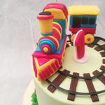 With a red, blue and yellow toy train running around a chocolate-y track on top of this toy train birthday cake for kids, the base of this design features a tall,green body with buttercream grass piped at the bottom, representing the open field through which a train might run.
